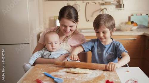 Young mother with little baby son looking at her older son rolling dough and cooking. Children cooking with parents, little chef, family having time together, domestic kitchen. photo