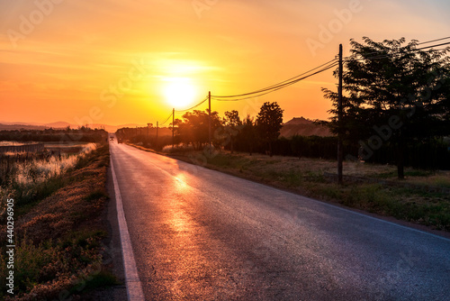 Landscape with a straight road with lamp posts on one side and vehicles circulating with the lights on at the end of the road, with the sunrise sun.