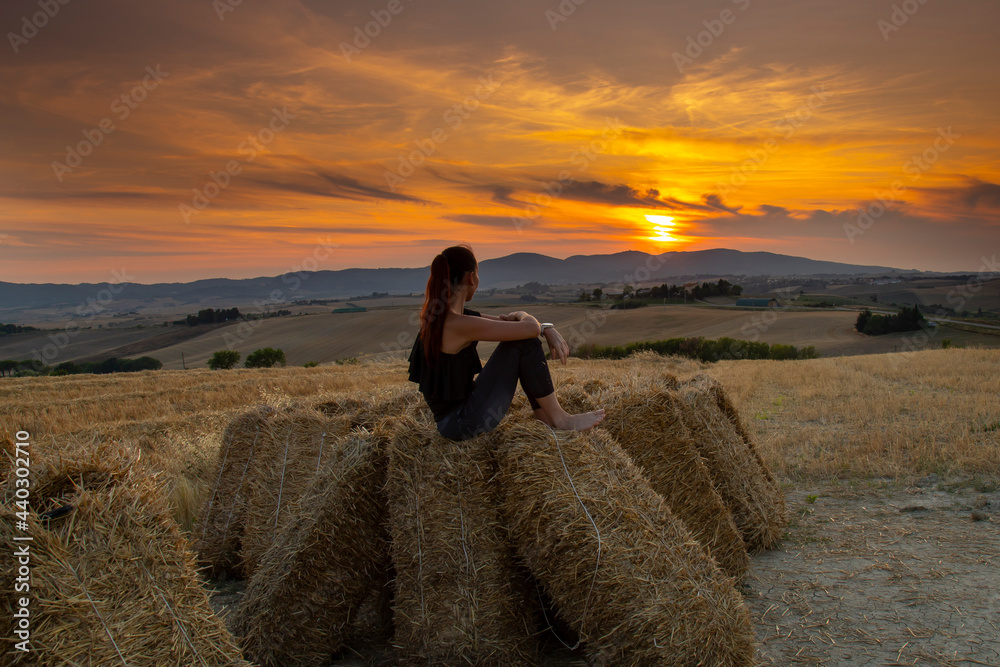 barefoot girl sits on a haystack and looks into the distance at the sunset