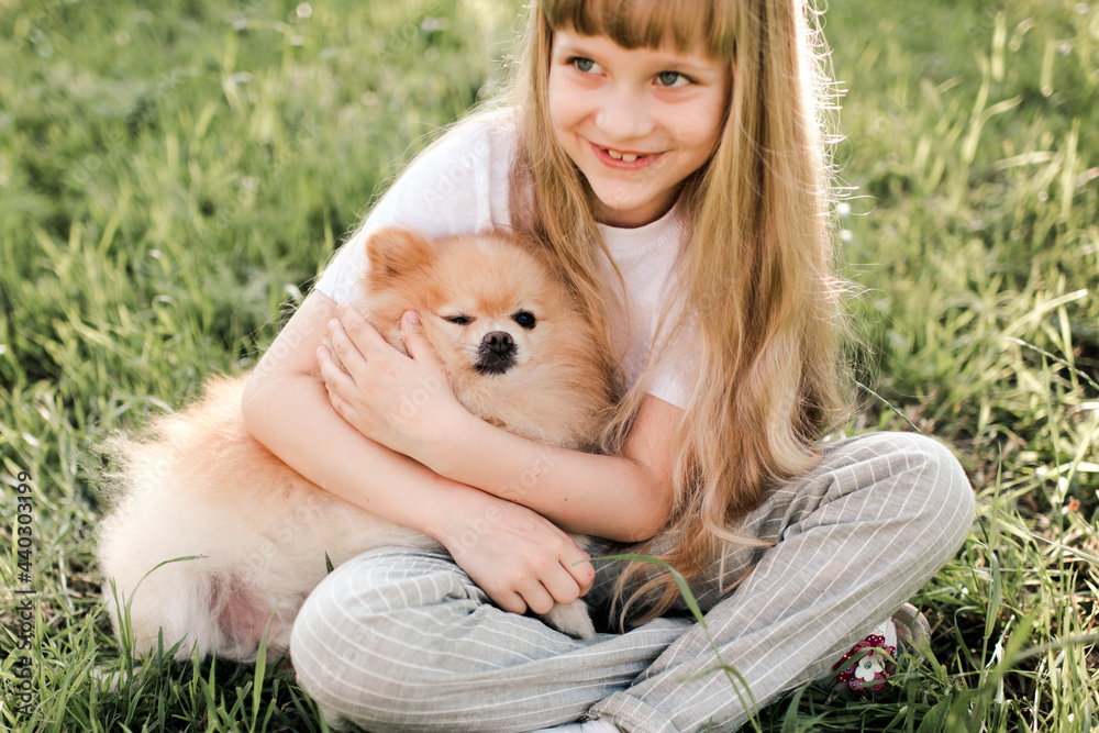 Pretty blonde kid girl 5-6 year old hold domestic pet sitting on green grass lawn outdoors in park. Looking at camera. Happiness. Friendship. Focus on dog. Pretty blonde kid girl 5-6 year old hold domestic pet sitting on green grass lawn outdoors in park. Looking at camera. Happiness. Friendship. Focus on dog.