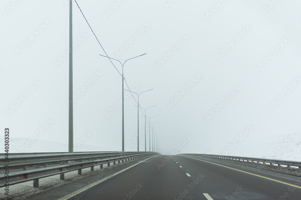 Road highway winter. A gray, bleak landscape with a wide highway without cars. The concept of bad weather, long-distance independent travel, self-isolation. Winter landscape in neutral colors. Fog