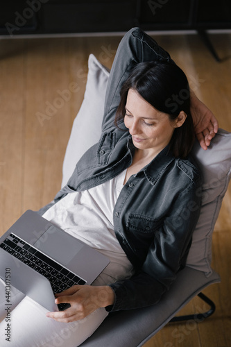 top view, European business woman in a gray shirt and white jeans sitting in a chair, working with a laptop and talking on the phone, texting, internet surfing or doing online shopping, online