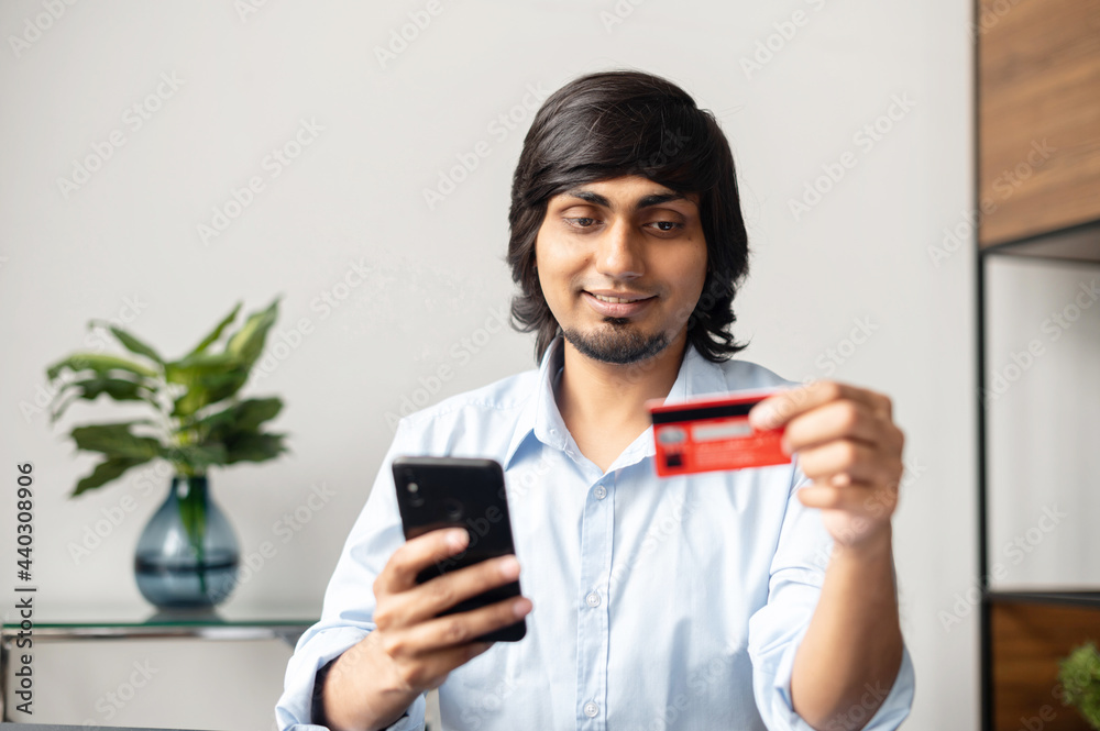Convenience to do shopping without leaving home. Young indian man holds smartphone and credit card, making purchases, ordering food delivery, inputting card details Convenience to do shopping without leaving home. Young indian man holds smartphone and credit card, making purchases, ordering food delivery, inputting card details