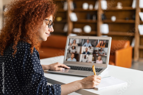 Female student with curly hairstyle using app for distance video communication, studying online, taking courses while staying at home, looking at laptop screen with group of people, taking notes