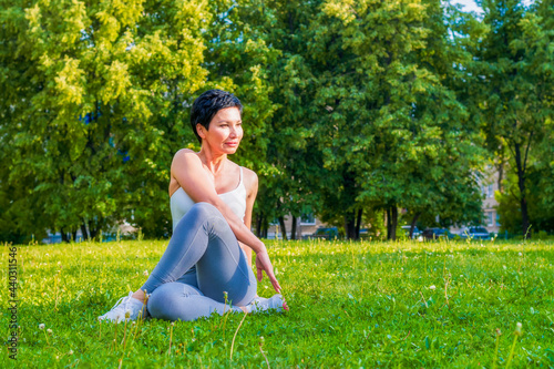 A middle aged brunette woman, dressed in a tracksuit, performs yoga exercises on a green stadium floor. Outdoor activities.