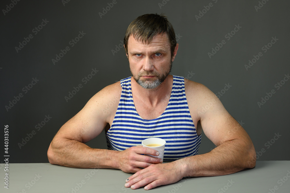 Portrait of an adult Caucasian serious man in a T-shirt drinking coffee at a table on a gray background. Portrait of an adult Caucasian serious man in a T-shirt drinking coffee at a table on a gray background.