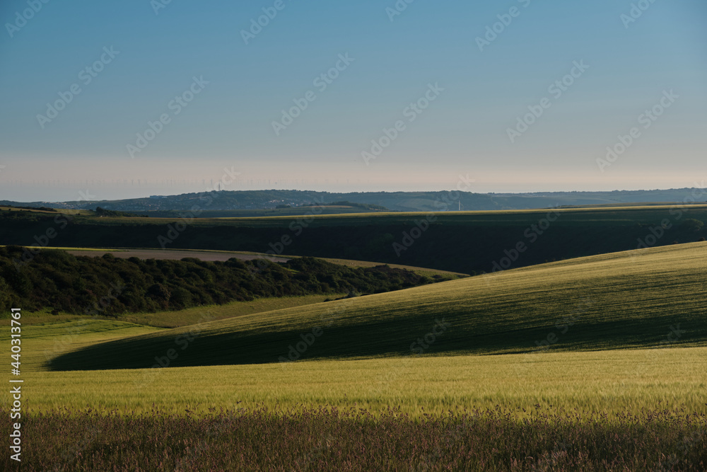 Farmland on a sunny evening in the South Downs National Park with the sun setting over the Sussex Weald. Newhaven and wind turbines in the far distance. 
