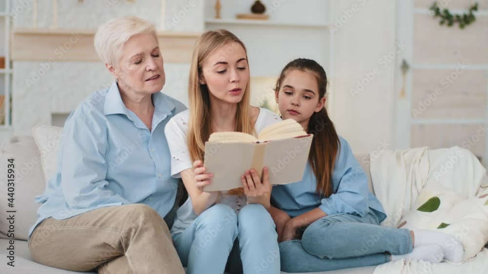 Headshot happy loving middle-aged woman and little daughter granddaughter listening grown up smiling daughter and mother reading paper fairy tale book. Female family sits on sofa spend home weekend Headshot happy loving middle-aged woman and little daughter granddaughter listening grown up smiling daughter and mother reading paper fairy tale book. Female family sits on sofa spend home weekend