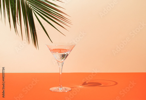 Top view of green tropical palm leaf and martini glass on orange and sand color background. Summer drink minimal concept. Elegant composition.