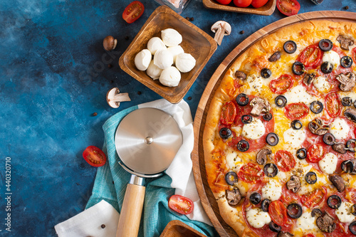 In the right corner of the photo We see a piece of pizza. On the left are a lot of ingredients - tomatoes, mushrooms, and a pizza knife. Dark blue background.