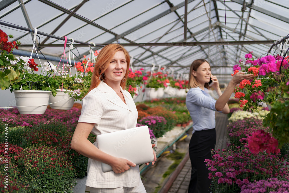 Businesswoman posing with a laptop while her partner discussing on the phone a proposal in a green house with flowers. Small business development concept.