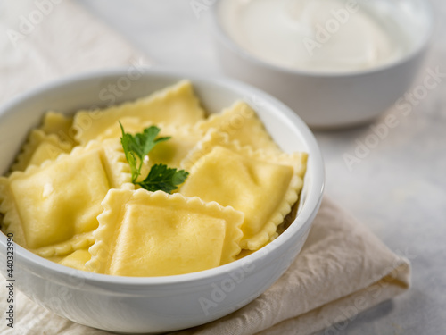 In the photo we see ravioli in a deep white bowl. In the upper right corner of the photo is a white bowl of sour cream. The ravioli is covered with a piece of green herbs. White napkin..