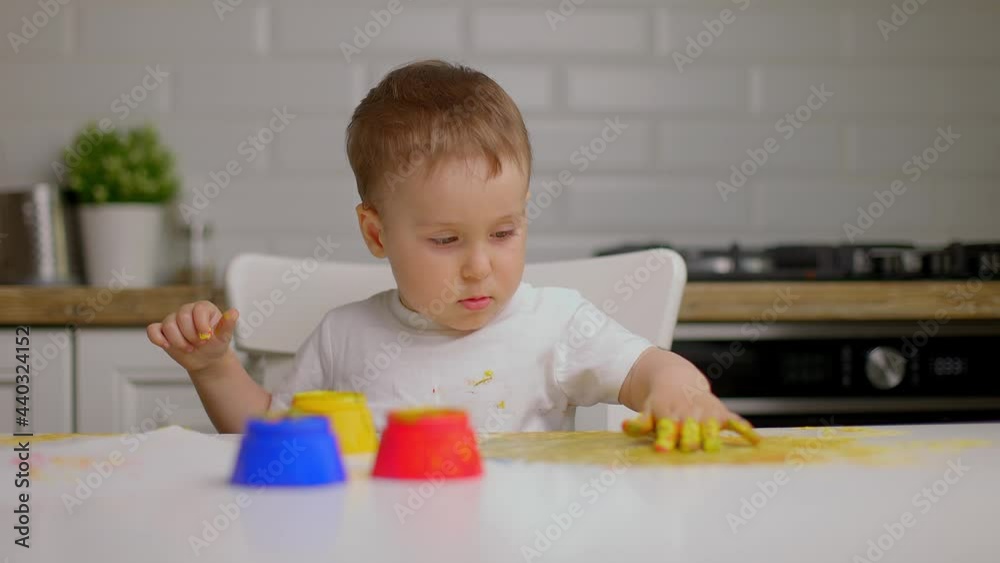 Handsome little baby boy draws with his hand on the kitchen table with finger paints. Slow motion Handsome little baby boy draws with his hand on the kitchen table with finger paints. Slow motion