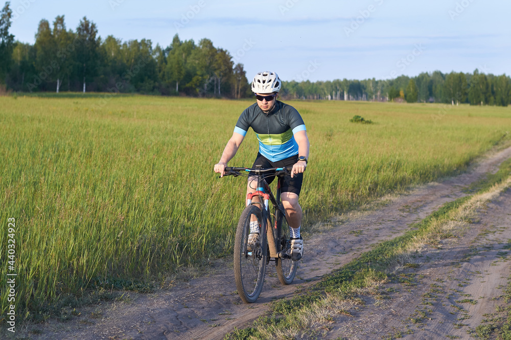 The guy is a cyclist leisurely rides a mountain bike on a dirt road. Dressed in a bicycle uniform, helmet and sunglasses. Endurance training. Healthy lifestyle and weight loss. Sunny weather.