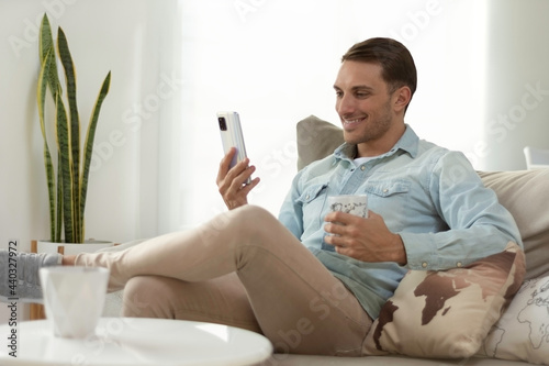 hombre joven mirando su telefono movil mientras toma un cafe sentado en el sofa de su casa photo