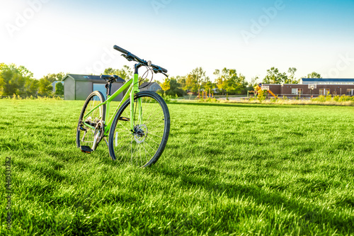 Bicycle parking at green field with blurred background of public school buildings and playground under sunny clear sky, idyllic nature outdoor. Relaxation activity in summer weekend, rural lifestyle.