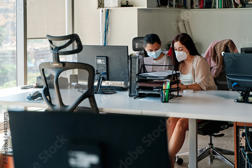 two young latina business women talking and working together in the office, wearing covid-19 protective masks in times of pandemic, teamwork concept