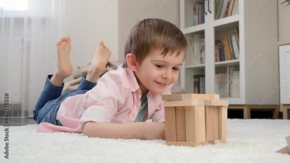Cute smiling boy lying on floor and looking at toy house he built from toy wooden blocks. Concept of child education and games at home Cute smiling boy lying on floor and looking at toy house he built from toy wooden blocks. Concept of child education and games at home