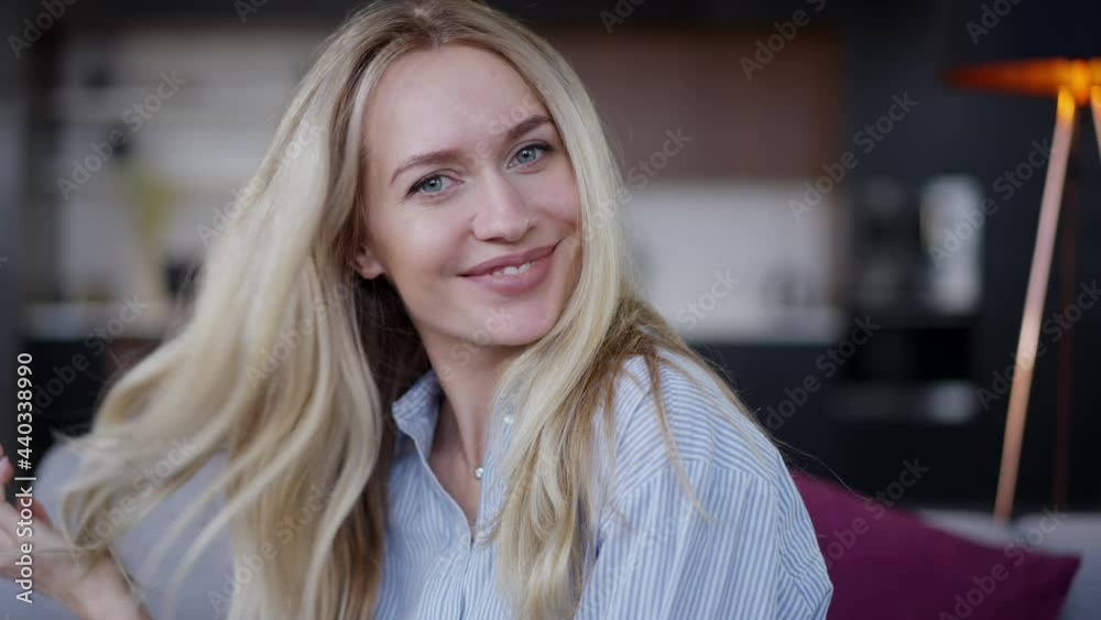 Close-up portrait of charming blond Caucasian young woman with blue eyes looking at camera smiling touching hair in slow motion. Positive happy relaxed millennial posing indoors at home in living room Close-up portrait of charming blond Caucasian young woman with blue eyes looking at camera smiling touching hair in slow motion. Positive happy relaxed millennial posing indoors at home in living room