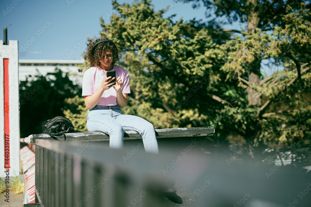 Fototapeta premium Cute young woman sitting outdoors with headphones on and choosing good music on her phone.