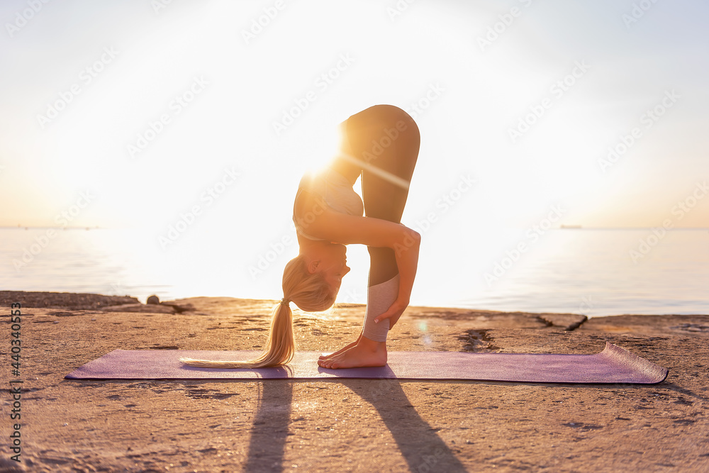 Young blonde woman in sportswear doing yoga asanas Uttanasana pose on the  seashore at sunrise on