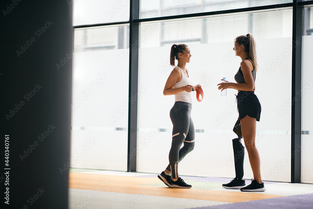 Fototapeta premium Happy female athletes talking while taking a break from exercising at gym. One of them has prosthetic leg.