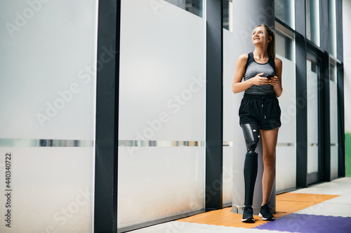 Happy athletic woman with prosthetic leg uses smartphone while taking a break during sports training at gym.