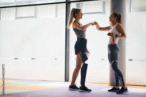 Fototapeta Naklejka Na Ścianę i Meble -  Happy sportswoman with leg disability and her female friend greeting with fists after successful training at gym.