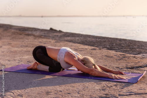 Young blonde woman in sportswear doing yoga asanas Balasana child pose on the seashore at sunriseon a purple yoga mat. Sports activities, meditation by the ocean on yellow sand photo