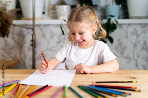 a little blonde girl is sitting at the table, smiling and drawing a heart with a red pencil, there are a lot of colored pencils on the table, learning concept