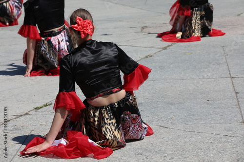 A female dancer in a red and black dress with a flower in her hair with a group of girls performs a dance sitting on the ground in a festive performance in a street square.Outdoor youth event