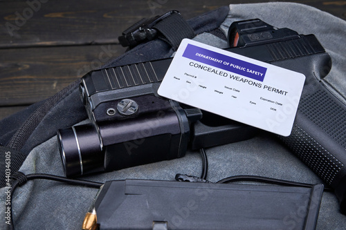 Handgun with red dot optic and weapon-mounted light laying on backpack on a table with spare magazine.