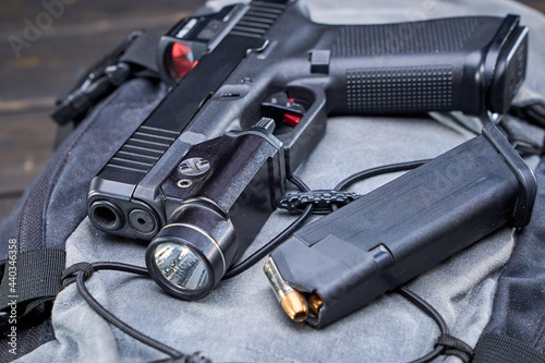 Handgun with red dot optic and weapon-mounted light laying on backpack on a table with spare magazine.
