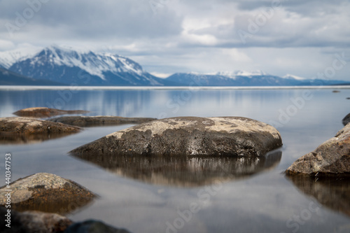 Scenic landscape in northern Canada during spring time with rocks, snow capped mountains reflection in calm lake water on cloudy afternoon.