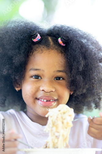 A little curly-haired African American girl sits at the table eating delicious Spaghetti Carbonara. Fun, cheerful. Appetite. Childhood and eating concepts.