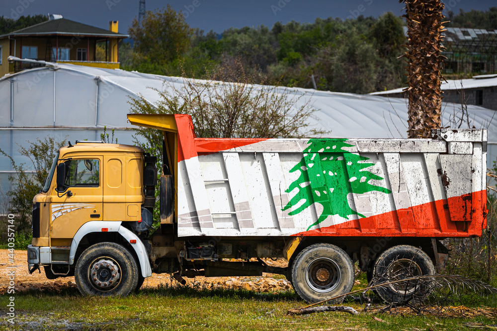 Dump truck with the image of the national flag of Lebanon is parked against the background of the countryside. The concept of export-import, transportation, national delivery of goods Dump truck with the image of the national flag of Lebanon is parked against the background of the countryside. The concept of export-import, transportation, national delivery of goods
