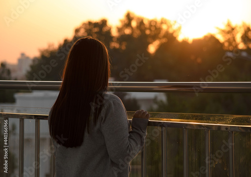Girl looks at the sunset among the trees, stands on a glass bridge, rear view from the back