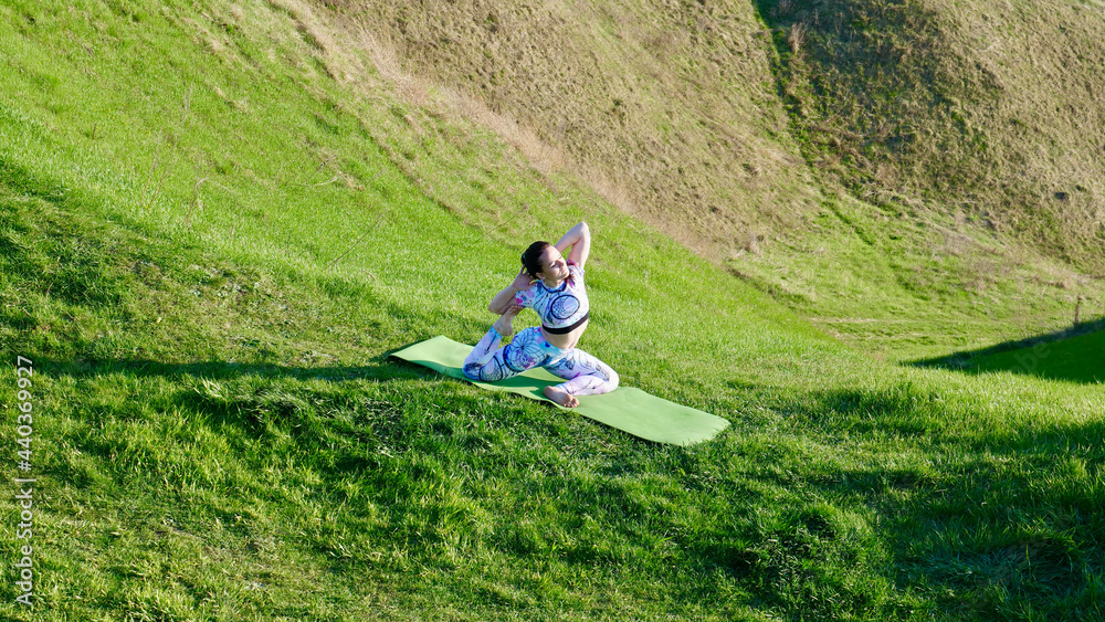 Woman yogi practicing yoga in nature, green hills in summer on a yoga mat. A brown-haired, Caucasian girl in an asana in sportswear, leggings and a T-shirt with braided hair in a ponytail.
