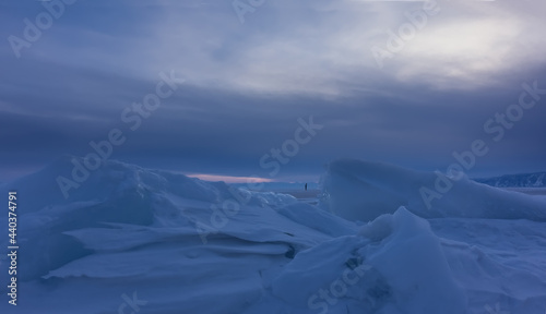 Snowdrifts and ice floes against the background of the evening cloudy sky. Close-up. In the distance, you can see the tiny silhouette of a lonely man walking on a frozen lake. Baikal