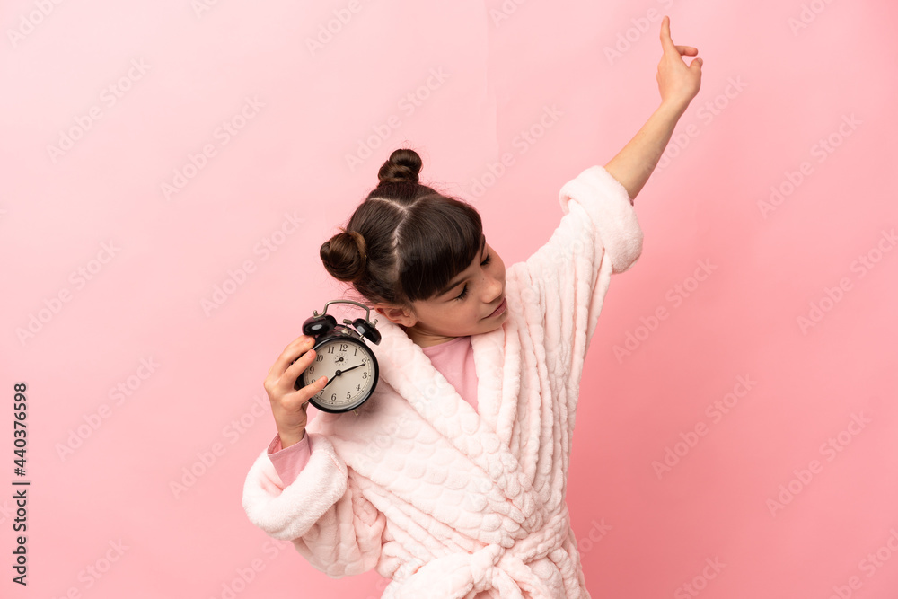 Little caucasian girl isolated on pink background in pajamas and holding clock with happy expression