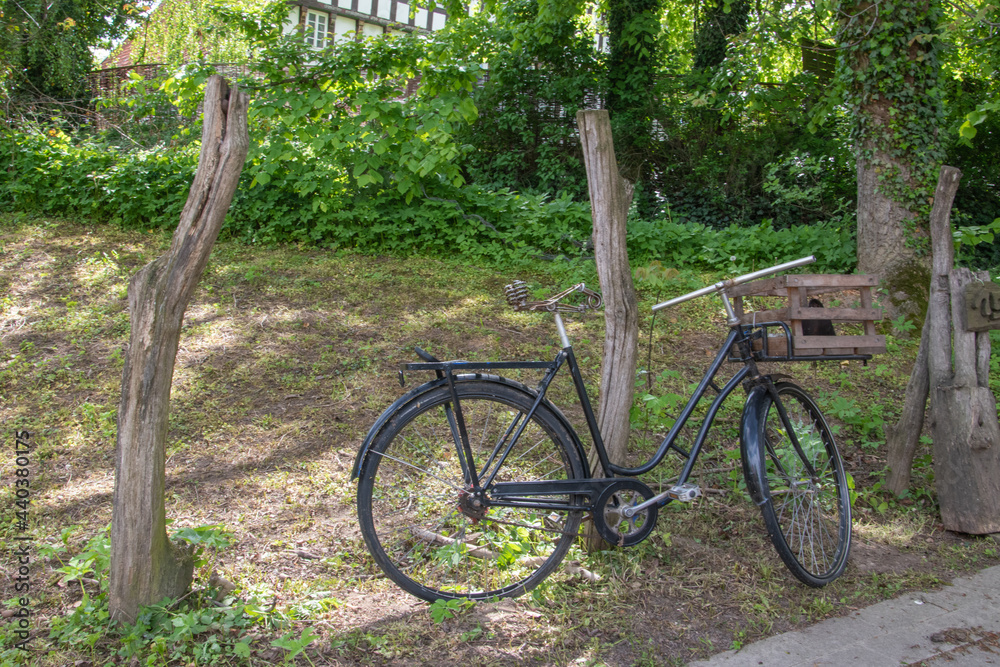 Altes schwarzes Fahrrad mit einem Holzkorb vor dem Lenker lehnt an einem Holzpfahl.