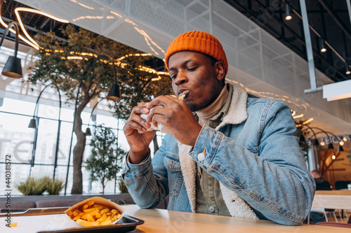 Fototapete Young black man in orange knitted hat eats tasty hamburger at table with french