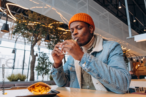 Young black man in orange knitted hat eats tasty hamburger at table with french fries on tray in decorated cafe close view