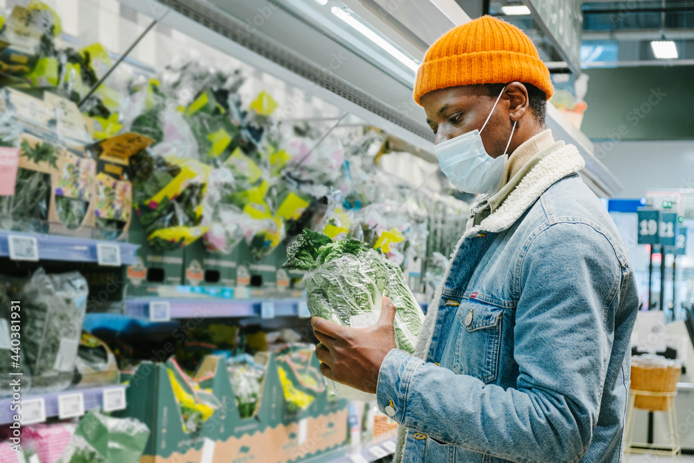 Black man shopper in orange hat with protective mask chooses Chinese cabbage near racks in supermarket department side view Black man shopper in orange hat with protective mask chooses Chinese cabbage near racks in supermarket department side view