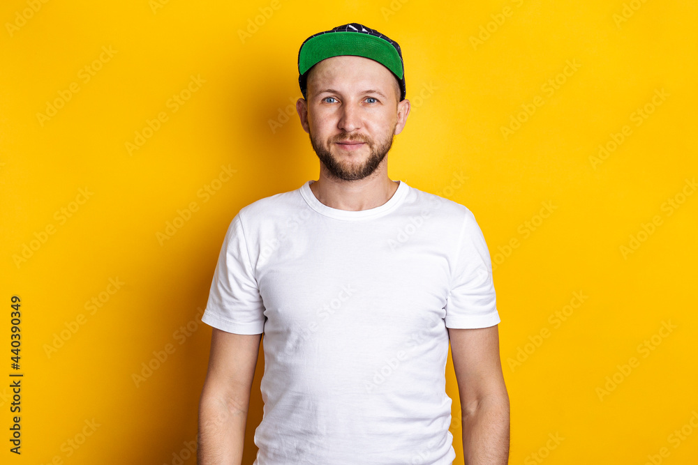 Bearded young man in a baseball cap and white T-shirt on a yellow background