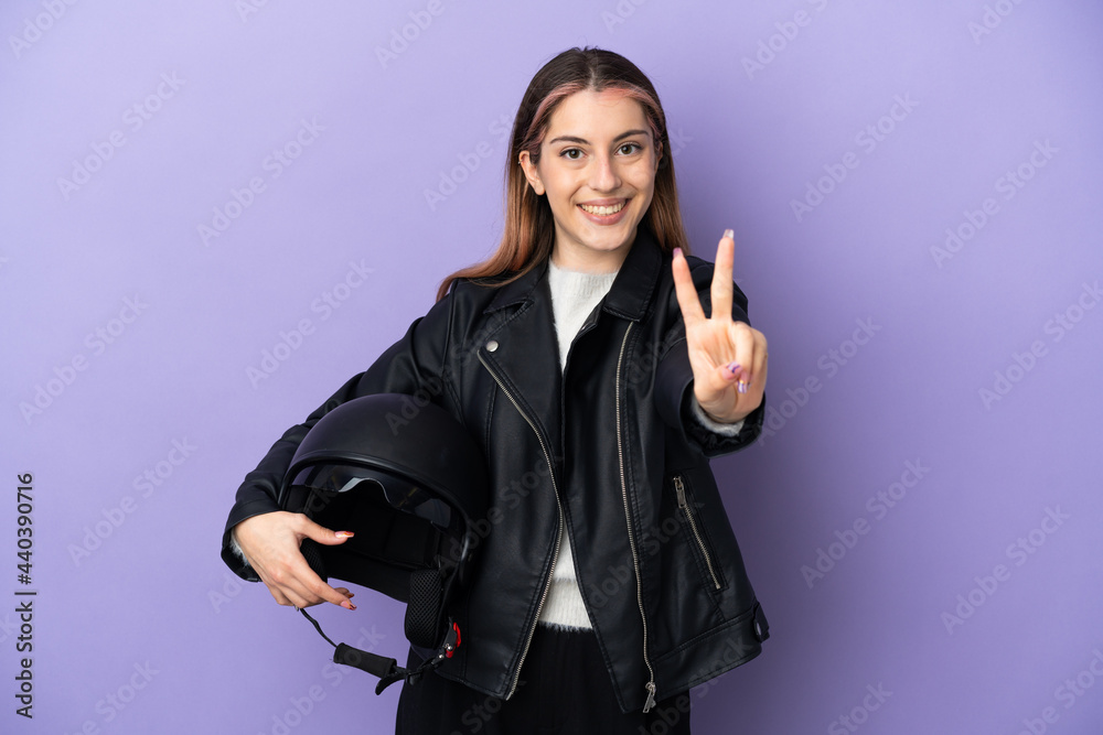 Young caucasian woman holding a motorcycle helmet isolated on purple background smiling and showing victory sign Young caucasian woman holding a motorcycle helmet isolated on purple background smiling and showing victory sign