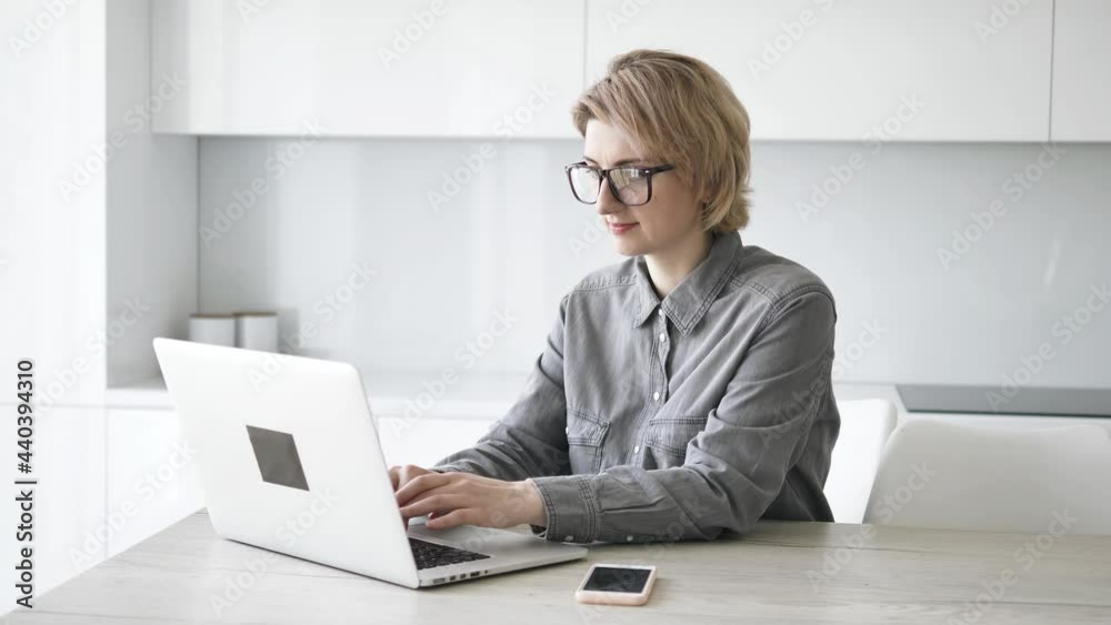 Short haired blonde businesswoman in grey shirt and glasses types on white laptop at wooden table with smartphone at home Short haired blonde businesswoman in grey shirt and glasses types on white laptop at wooden table with smartphone at home
