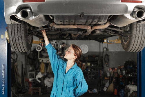 A girl car mechanic with glasses in the garage checks the fastening of the wheels and the integrity of the chassis in a car workshop. Car repair in the garage. Woman in work clothes.