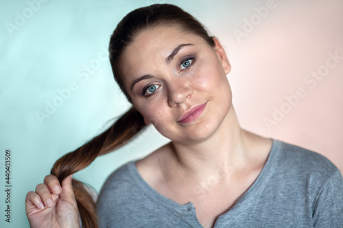 Closeup portrait of young brunette woman with blue eyes, smiling and curling her pony tail on vibrant cyan and pink background. Natural make-up, round shape face, body positive concept. Copy space.