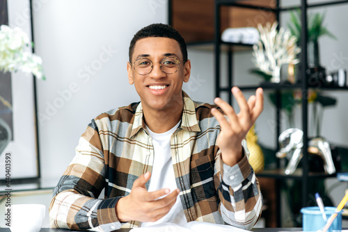 Video conference. Handsome friendly latino guy in glasses and in a casual shirt, sits at his work desk, looks at the camera, communicates on a video call, gesturing with hands, smiles pleasant